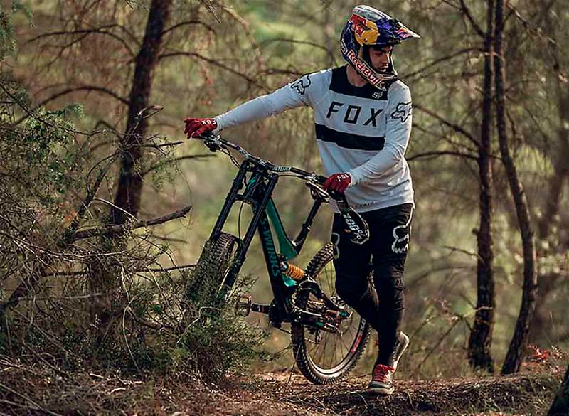 Ciclista em local arborizado segurando sua bicicleta e utilizando os equipamentos para a prática de downhill 